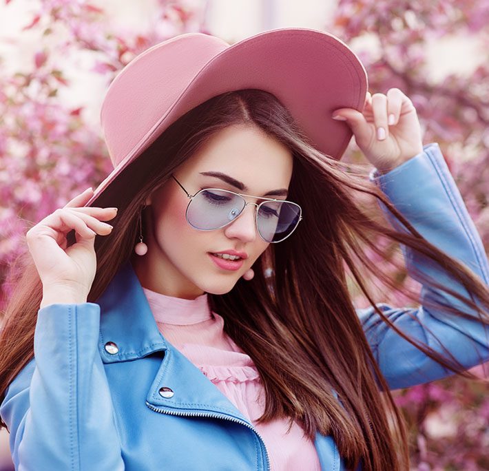 Junge Frau mit rosa Schlapphut und blau getönter Sonnenbrille vor rosa blühendem Strauch